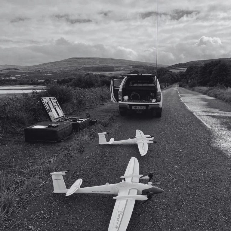 Two fixed-wing drones on a gravel road beside a parked vehicle and a scenic landscape ready to perform a topographic survey in Scotland.