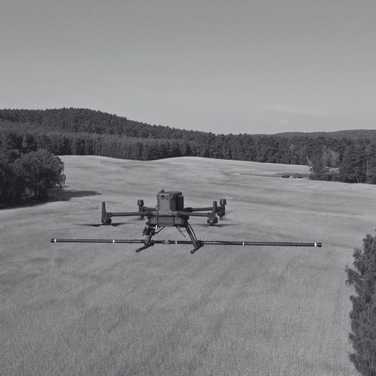 A drone flying over a vast, open field with trees in the background conducting a magnetometer survey for sub-surface anomalies